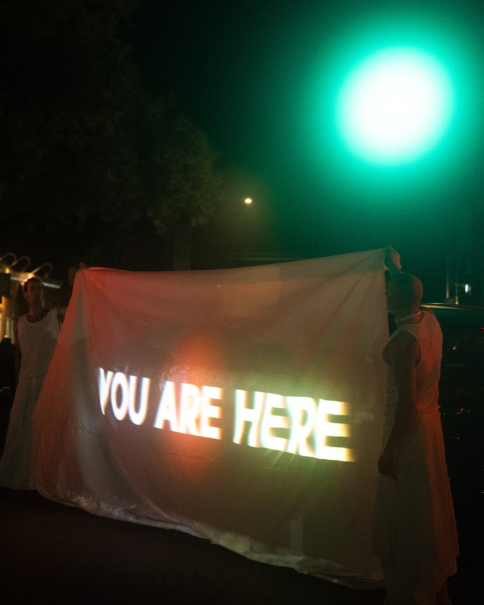 Performers holding up white fabric with the words you are here projected on it